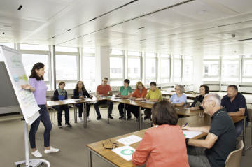Eine Vortragende spricht an der Flipchart vor mehreren Kursteilnehmer*innen, die an Tischen sitzen. 